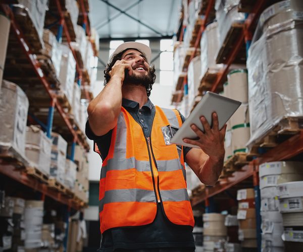 A man talking on a phone while walking through a warehouse with his tablet.